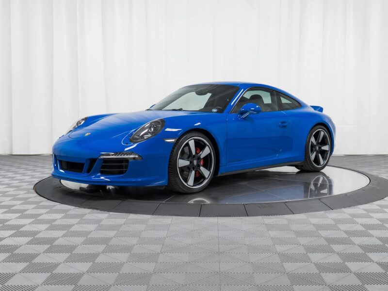 A blue Porsche 911 sports car—one of the best manual transmission cars—is displayed on a circular platform in a showroom with a white curtain backdrop and checkered floor.