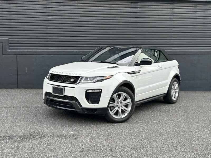 A luxury SUV, this white Range Rover Evoque convertible is parked on a paved surface in front of a dark gray metal wall.