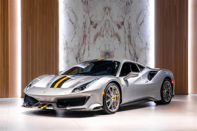 A track-ready silver Ferrari 488 Pista with yellow and black racing stripes is parked indoors against a modern backdrop with wood paneling and a marble wall.