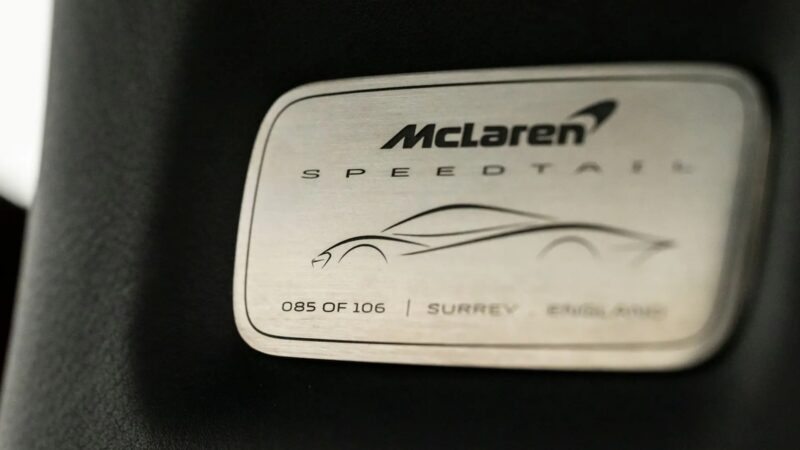 Close-up of a McLaren Speedtail plaque indicating this ultra-low mile Speedtail, number 85 of 106 with $250k in MSO options, is heading to auction, featuring a car outline and "McLaren Speedtail" engraved.