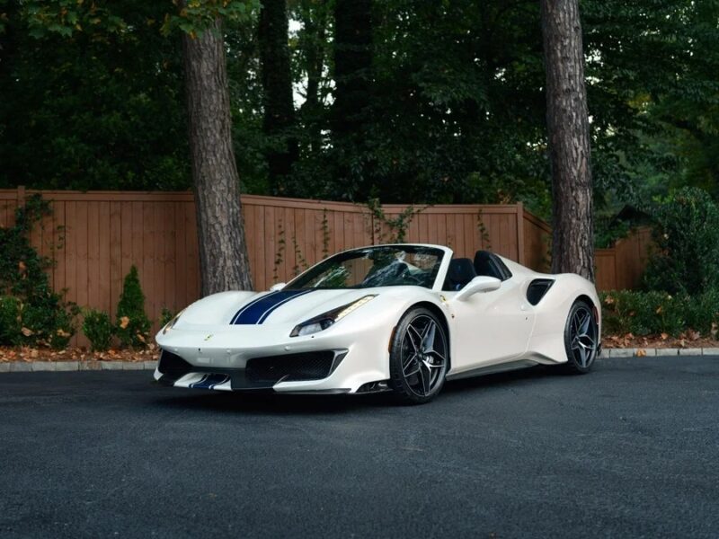 A white Ferrari 488 Pista convertible with blue racing stripes is parked on a paved surface in front of a wooden fence and trees, highlighting its track-ready Ferrari spirit.