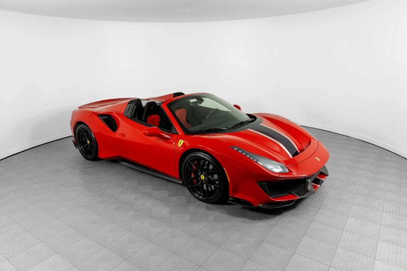 A red Ferrari 488 Pista convertible sports car with black racing stripes, track-ready and pristine, is parked in a white, curved photo studio with a gray tiled floor.