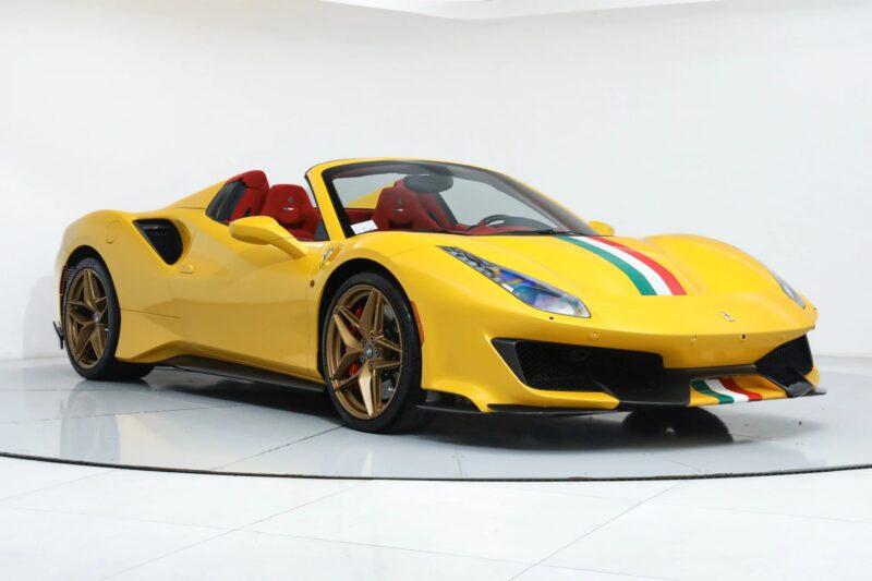 A yellow Ferrari 488 Pista with red interior, gold wheels, and green, white, and red racing stripes is displayed in a white showroom, highlighting this stunning track-ready Ferrari for sale.