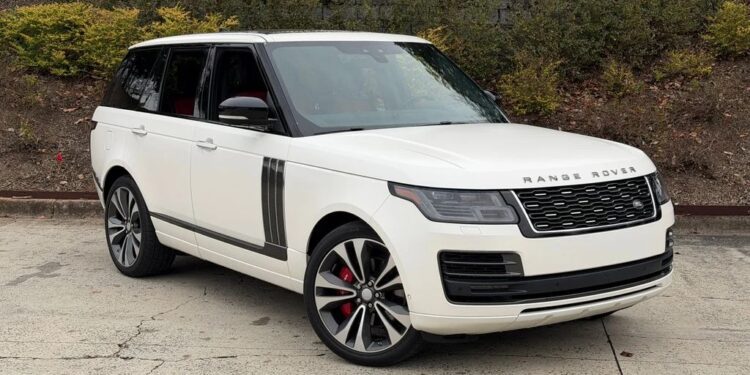 A white Range Rover SUV, one of the best luxury SUVs, is parked on a concrete surface beside a planted embankment with bushes and a metal fence above.