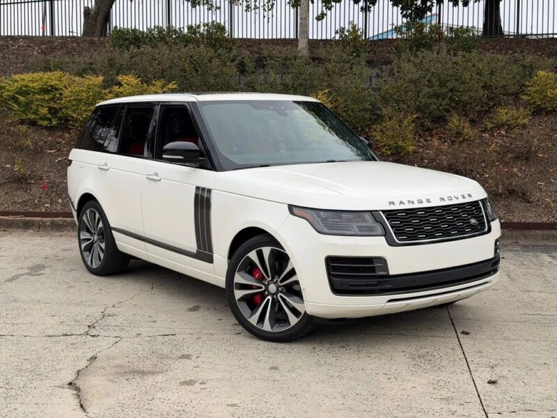 A white Range Rover SUV, one of the best luxury SUVs, is parked on a concrete surface beside a planted embankment with bushes and a metal fence above.