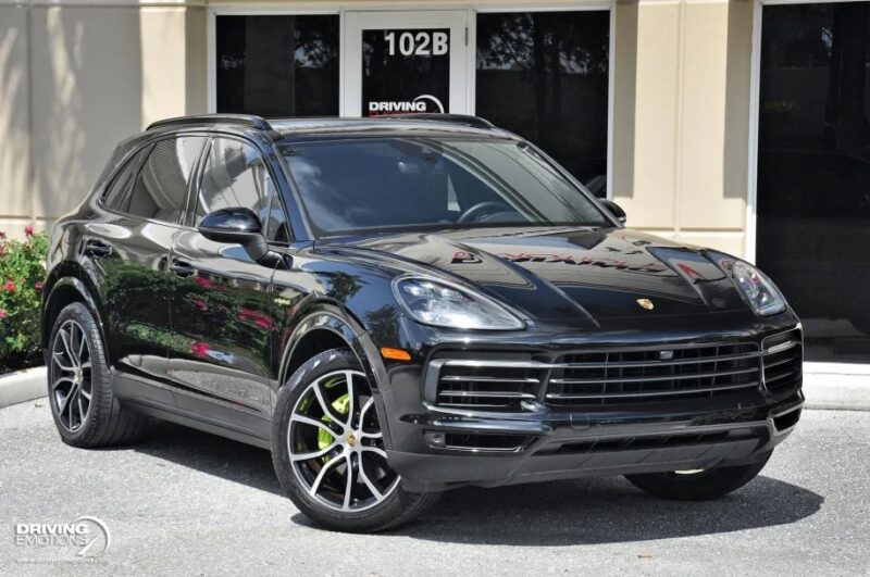 A black Porsche Cayenne luxury car is parked in front of a beige commercial building with glass doors and windows, perfect for those seeking high-end cars for sale.