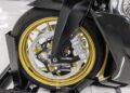 Close-up of a 2022 Aston Martin AMB 001 motorcycle front wheel with a gold rim, disc brake, and black carbon fiber fender, displayed on a stand in a well-lit indoor setting at Sotheby's Motorsport.