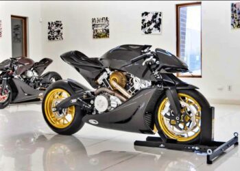 A sleek black Aston Martin AMB 001 motorcycle with gold wheels is displayed on a stand inside a modern showroom with white walls, windows, and art. Another motorcycle is in the background at Sotheby’s Motorsport in 2022.
