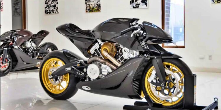 A sleek black Aston Martin AMB 001 motorcycle with gold wheels is displayed on a stand inside a modern showroom with white walls, windows, and art. Another motorcycle is in the background at Sotheby’s Motorsport in 2022.