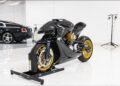 A 2022 Aston Martin AMB 001 motorcycle with gold wheels is displayed on a stand in a white showroom; a black luxury car is parked in the background.