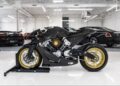 A black AMB 001 motorcycle with gold rims is displayed on a stand in a bright showroom, with two black luxury cars—including an Aston Martin—in the background.
