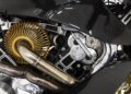 Close-up of a 2022 Aston Martin AMB 001 motorcycle engine, featuring metallic pipes, gold-colored components, and black carbon fiber body panels, showcased by Sotheby's Motorsport.