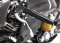Close-up of an Aston Martin AMB 001 motorcycle handlebar with a silver brake lever, carbon fiber body panel, and a yellow engine component visible in the background.