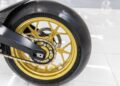 Close-up of a 2022 Aston Martin AMB 001 motorcycle's rear wheel, featuring a gold alloy rim, chain drive, and Michelin Power Slick tire on a shiny white tiled floor at Sotheby's Motorsport.
