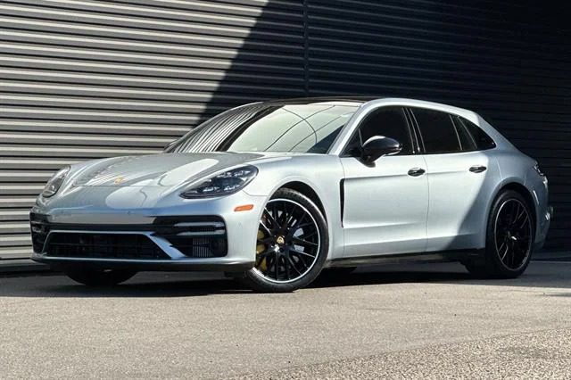 A silver Porsche Panamera, one of the finest Luxury Performance Wagons, is parked on a paved surface in front of a large, corrugated metal wall.