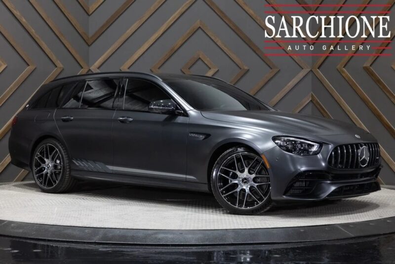A matte gray Mercedes-Benz AMG station wagon, part of the Luxury Performance Wagons collection, is parked indoors on a display platform, with geometric patterned walls and the Sarchione Auto Gallery logo in the background.
