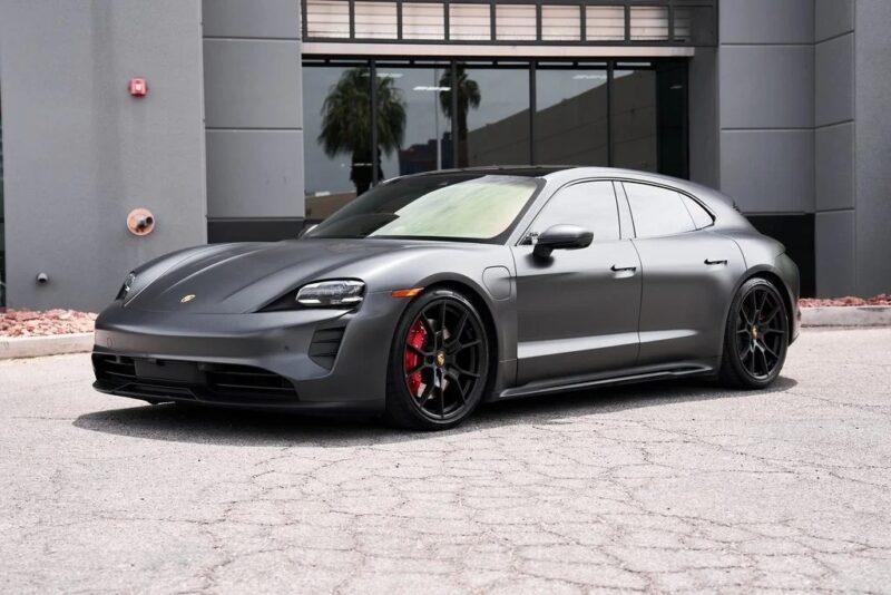 A matte black Porsche Taycan Cross Turismo, one of the most striking performance wagons, is parked in front of a modern gray building on a sunny day.