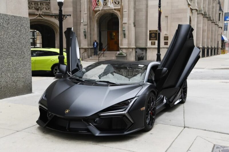A matte black Lamborghini luxury car with scissor doors open, parked on a city sidewalk in front of a stone building.