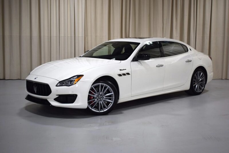 A white Maserati Quattroporte luxury sedan, a true icon of Italian luxury, is parked indoors on a light gray floor with beige curtains in the background.