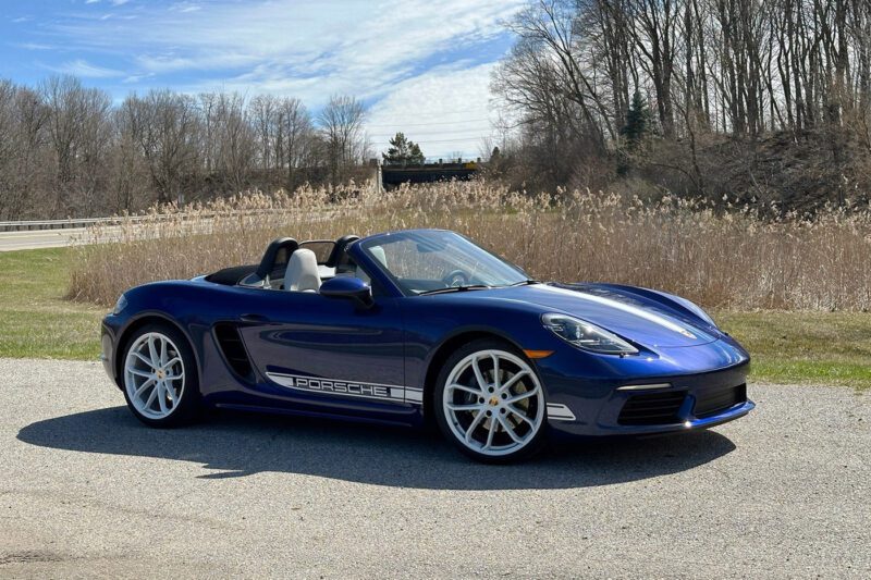 A blue Porsche convertible sports car, ideal for those seeking the best lease deals, is parked on a paved surface with dry grass, leafless trees, and a partly cloudy sky in the background.