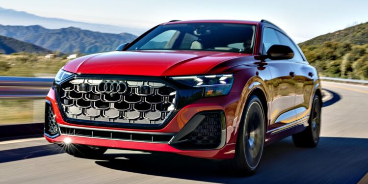 A red 2025 Audi SQ8 SUV drives on a winding road with mountains and trees in the background, showcasing its impressive style under a clear sky.