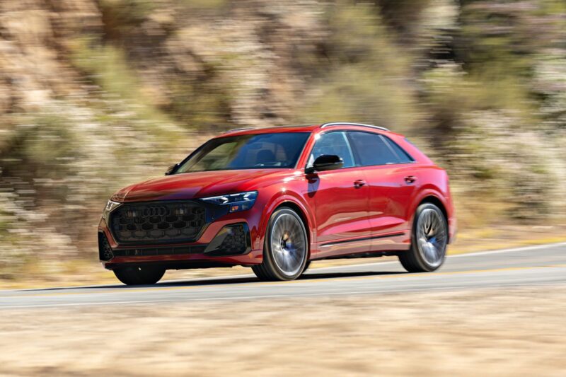 A red 2025 Audi SQ8 SUV is driving on a paved road with blurred trees and rocky hills in the background.