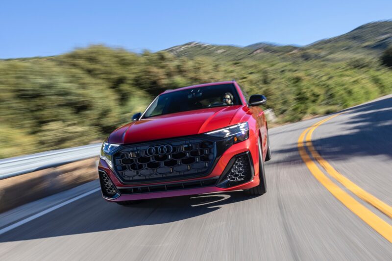 A red 2025 Audi SQ8 SUV drives on a curved mountain road with greenery and blue sky in the background.