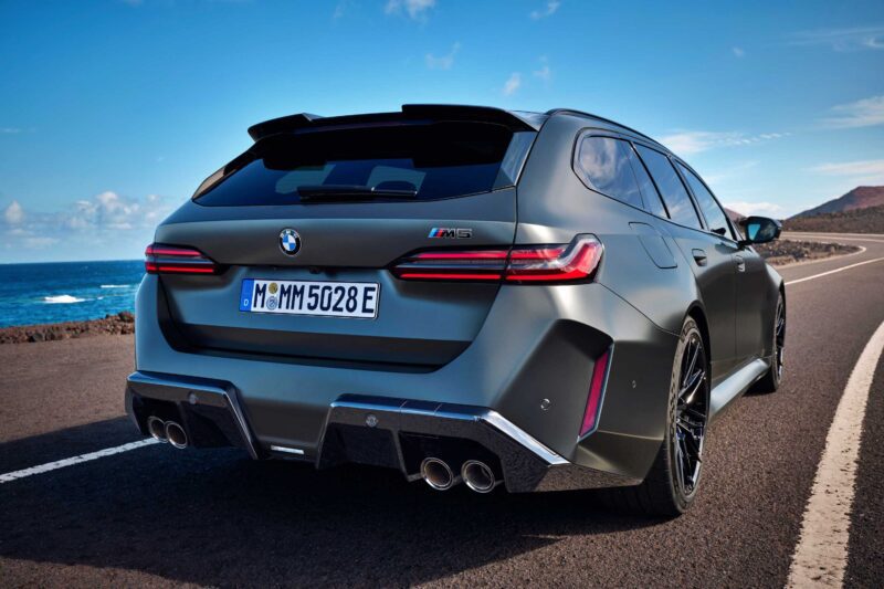 A matte grey BMW M5 Touring is parked on a coastal road with ocean and blue sky in the background, viewed from the rear—Review: 2025 BMW M5 Touring weighs in as a great allrounder.