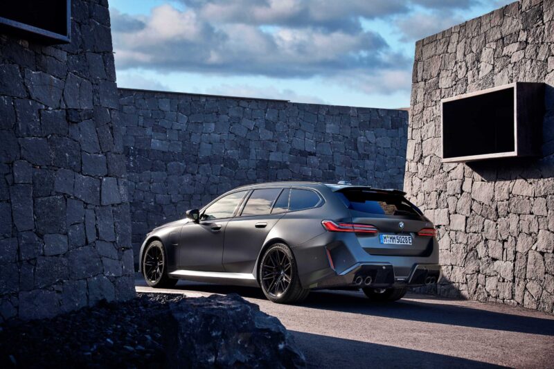 A matte gray BMW station wagon, seen from the rear three-quarter angle and parked between stone walls under a partly cloudy sky, hints at the versatility praised in Review: 2025 BMW M5 Touring Weighs In As A Great Allrounder.