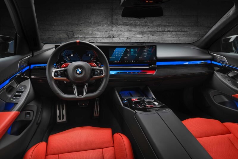 BMW car interior featuring a digital dashboard, a large touchscreen display, red leather seats, ambient blue lighting, and a modern steering wheel—true to the Review: 2025 BMW M5 Touring Weighs In As A Great Allrounder.