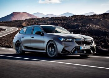 A matte gray BMW station wagon drives on a winding road through a mountainous, rocky landscape under a clear sky—Review: 2025 BMW M5 Touring weighs in as a great allrounder for any adventure.