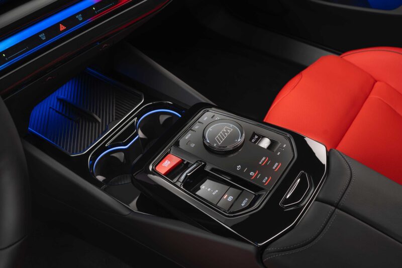 Close-up of a car’s center console featuring a gear selector dial, control buttons, cup holders, and black and red seats—echoing the refined interior seen in the Review: 2025 BMW M5 Touring Weighs In As A Great Allrounder.