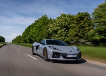A silver sports car drives down an empty rural road lined with green trees under a blue sky.