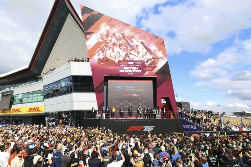 A large crowd gathers in front of the podium at the British GP, with drivers and officials on stage and a race-themed building in the background, celebrating moments like McLaren’s historic achievements at the F1 Diamond Jubilee.