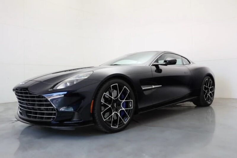 A sleek black luxury car is parked indoors on a polished concrete floor, shown from a front side angle against a plain white background.
