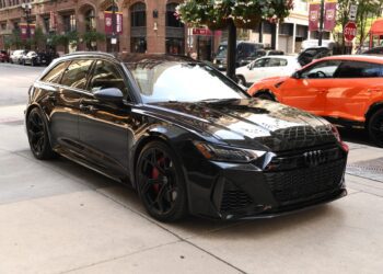 A black Audi RS6 Avant, one of the top luxury performance wagons, is parked on a city street, with buildings, signs, and an orange car in the background.