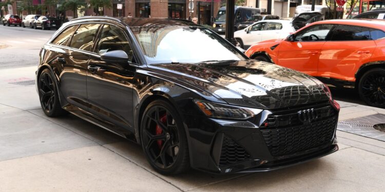 A black Audi RS6 Avant, one of the top luxury performance wagons, is parked on a city street, with buildings, signs, and an orange car in the background.