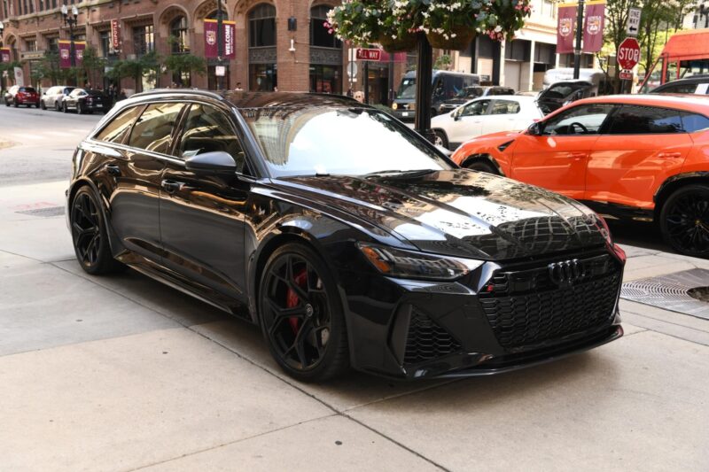 A black Audi RS6 Avant, one of the top luxury performance wagons, is parked on a city street, with buildings, signs, and an orange car in the background.