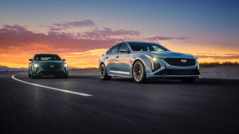 Two modern sedans, including the last V8-powered sedan to offer a manual in 2025, drive along a curved road at sunset, headlights cutting through the scenic evening sky.