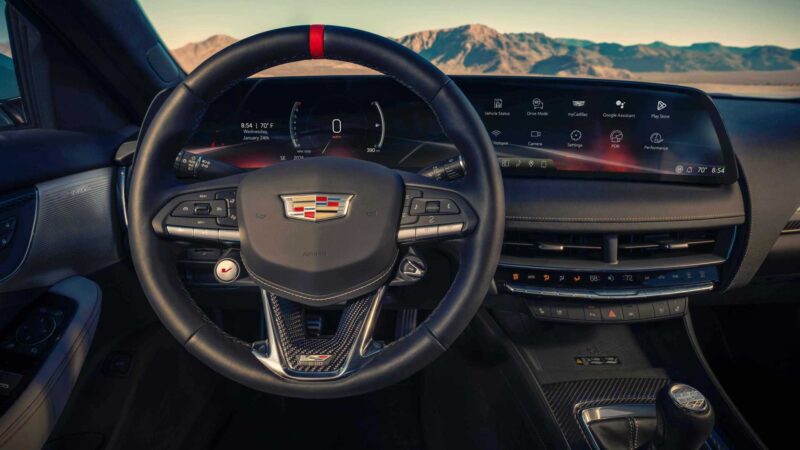 View of a Cadillac steering wheel and digital dashboard with mountain scenery visible through the windshield—the last V8-powered sedan to offer a manual in 2025.
