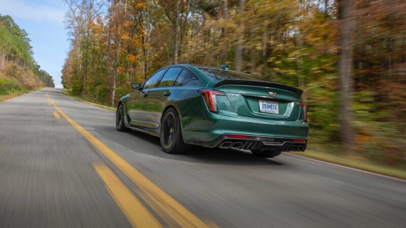 A green Cadillac sedan, the last V8-powered sedan to offer a manual in 2025, drives down a two-lane road lined with trees showing autumn foliage.