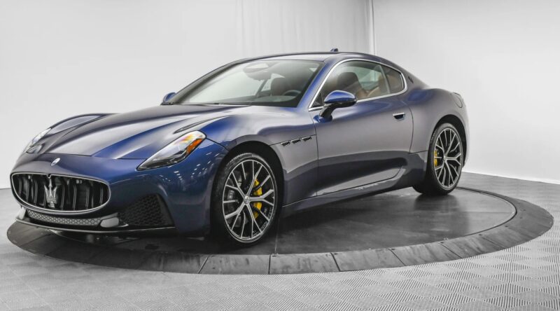A blue Maserati for sale, this Italian luxury sports coupe is parked on a rotating platform in a pristine white indoor showroom.