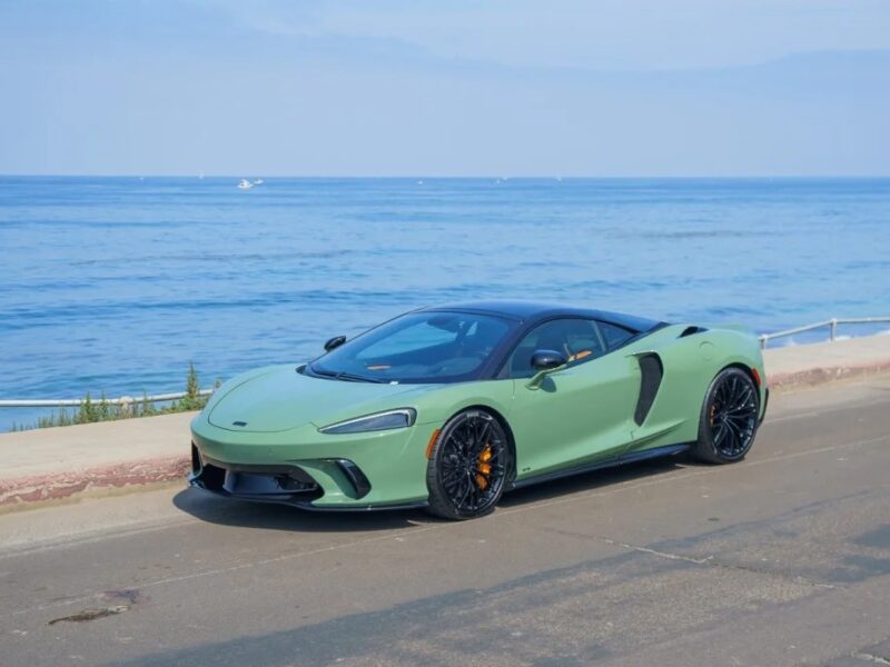 An exotic green sports car is parked on a coastal road, with the ocean and a blue sky in the background.