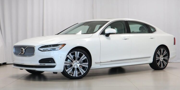 A white Volvo S90 sedan, one of the top affordable luxury sedans, is parked indoors on a tiled floor with a plain white background.