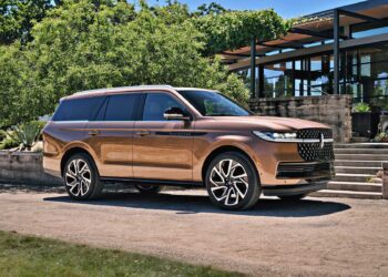 A bronze Lincoln Navigator SUV is parked on a gravel driveway in front of modern steps and greenery on a sunny day, echoing the comfort described in the Review: 2025 Lincoln Navigator L Black Label Coddles Like A Spa On Wheels.