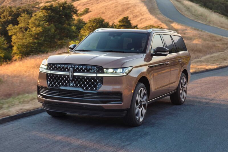 A brown Lincoln Navigator SUV drives on a paved road through a hilly landscape with dry grass and trees, embodying the luxury described in the review: 2025 Lincoln Navigator L Black Label Coddles Like A Spa On Wheels.