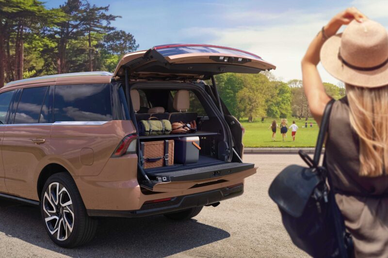 A woman in a hat stands near an SUV with the trunk open, packed with bags and outdoor gear, in a park with people in the background—like a scene from a Review: 2025 Lincoln Navigator L Black Label Coddles Like A Spa On Wheels.