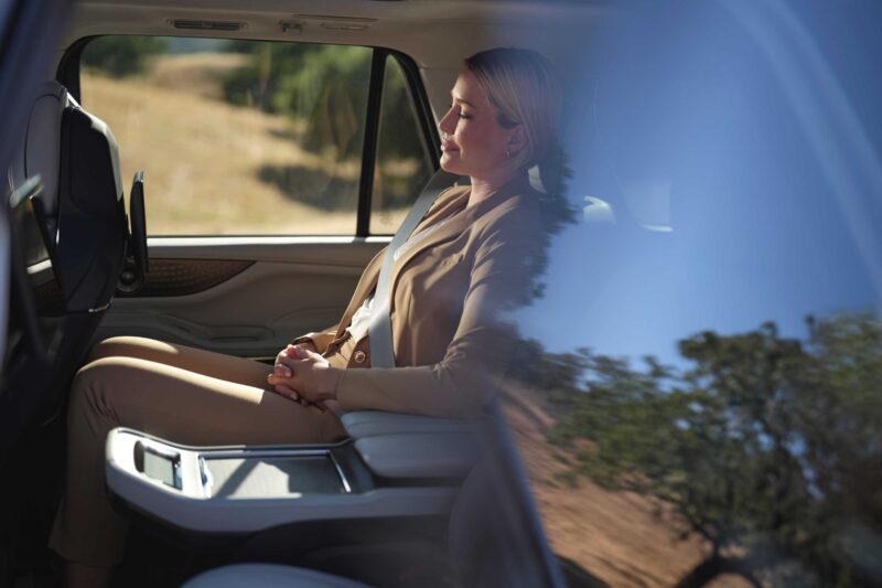 A woman in a tan suit sits in the back seat of a car, resting with her eyes closed; trees and landscape are visible outside the window—much like relaxing in the 2025 Lincoln Navigator L Black Label, which coddles like a spa on wheels.
