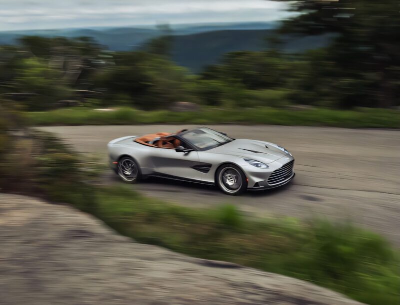 A silver 2026 Aston Martin Vanquish convertible with a tan interior and powerful V12 engine speeds along a curving road surrounded by lush greenery.