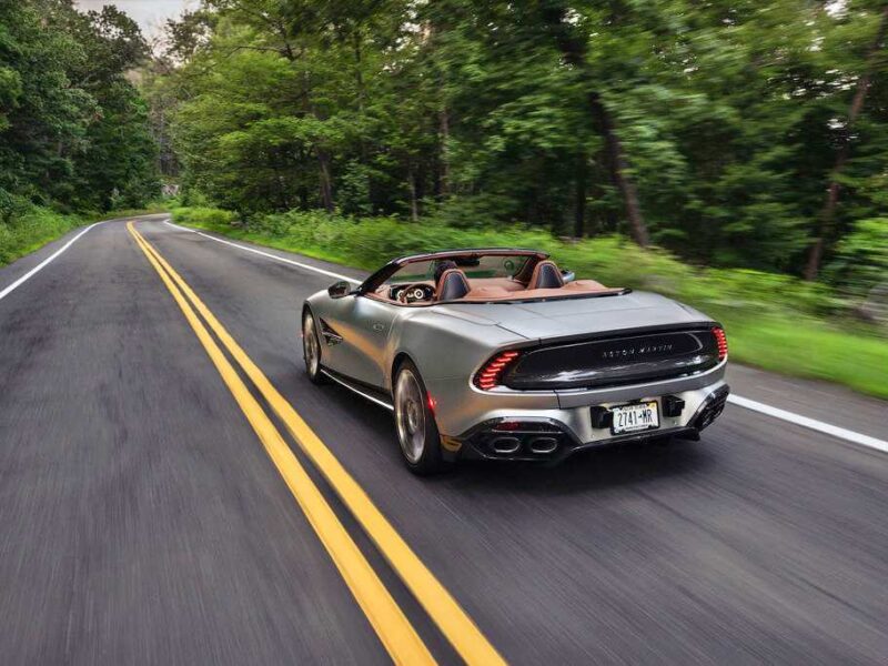 A silver 2026 Aston Martin Vanquish convertible sports car drives down a two-lane road surrounded by trees, its V12 engine purring and the top down, with two people inside.